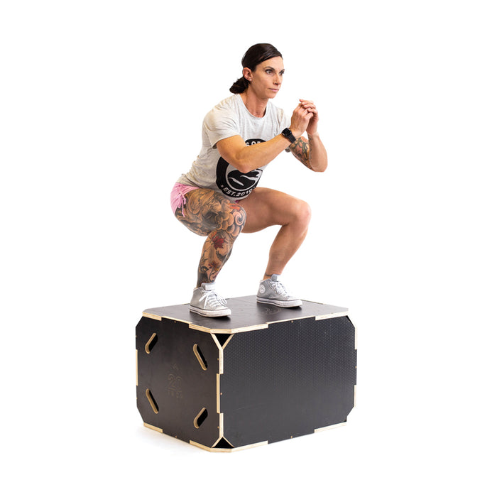 A woman with tattoos squats on a Bells of Steel 3 in 1 Anti-Slip Wood Plyo Box, wearing a light t-shirt, pink shorts, and gray sneakers, demonstrating essential home gym equipment against a white background.