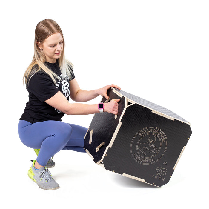 A woman in a black shirt, blue leggings, and gray sneakers kneels to assemble a Bells of Steel 3 in 1 Anti-Slip Wood Plyo Box, a large black gym essential marked "18 inch" with the brand logo on its side.