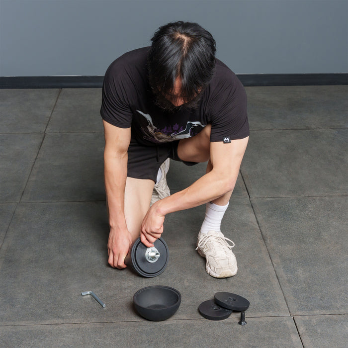 A person kneels on a tiled floor, assembling a dumbbell with black weights and a silver handle. Within reach are extra weights—including the Bells of Steel 6-12KG Adjustable Kettlebell—along with a tool, ready for use.
