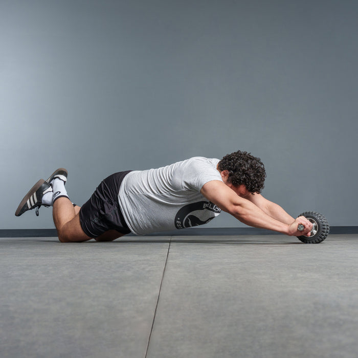 A man in a gray t-shirt and black shorts uses the Bells of Steel USA ABsyidyan Stainless Steel Ab Roller, kneeling on a gym floor as he performs an ab rollout with outstretched arms against a plain background.