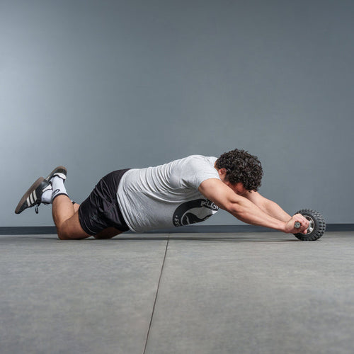 A man in a gray t-shirt and black shorts uses the Bells of Steel USA ABsyidyan Stainless Steel Ab Roller, kneeling on a gym floor as he performs an ab rollout with outstretched arms against a plain background.