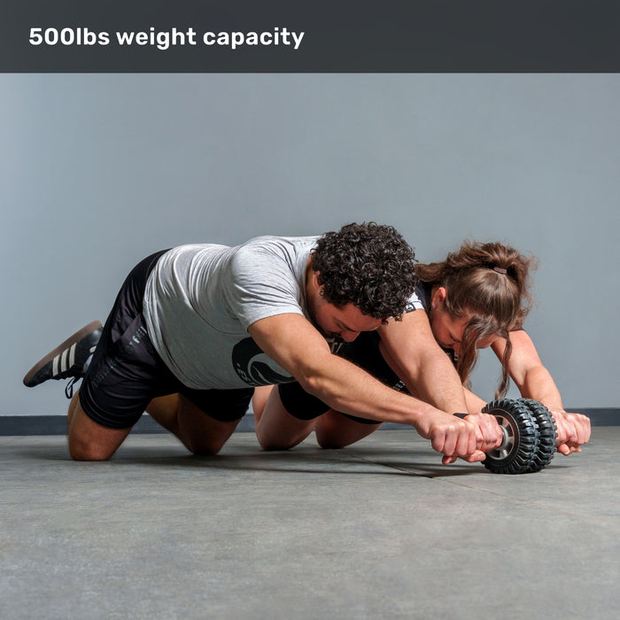 A man and woman kneel on the floor using the ABsyidyan Stainless Steel Ab Roller by Bells of Steel USA together. "500lbs weight capacity" appears at the top. The background is plain gray.