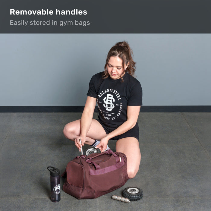A woman kneels, packing the ABsyidyan Stainless Steel Ab Roller by Bells of Steel USA—with removable handles and a dual wide wheel—into her gym bag. Text: “Removable handles—Easily stored in gym bags.”.