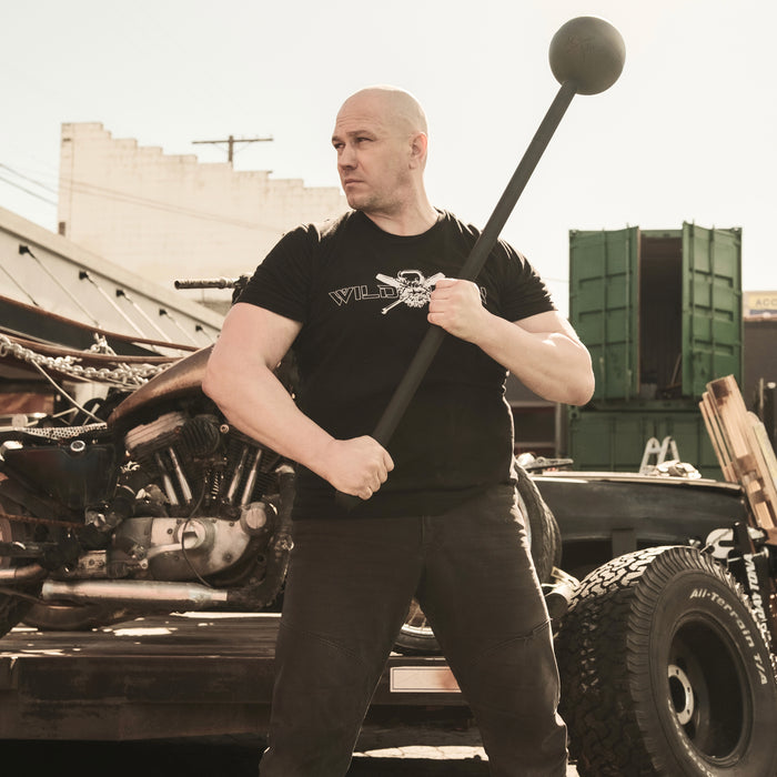 A muscular man in a black T-shirt holds his Bells of Steel Adjustable Macebell beside a motorcycle on a flatbed trailer. With a serious and focused demeanor, he's set to enhance his shoulder mobility and core strength, standing against an industrial area backdrop under broad daylight.