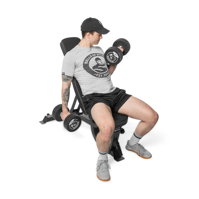 A man in a cap, gray t-shirt, black shorts, and athletic shoes uses the Bells of Steel Adjustable Utility Bench set to an incline to perform seated bicep curls with dumbbells.