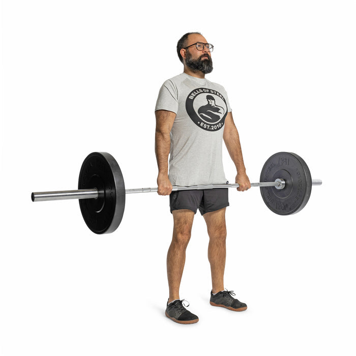 A bearded man in glasses, a gray T-shirt, black shorts, and sneakers stands in a lifting pose against a white background, holding a barbell loaded with Bells of Steel All-Black Bumper Plates.