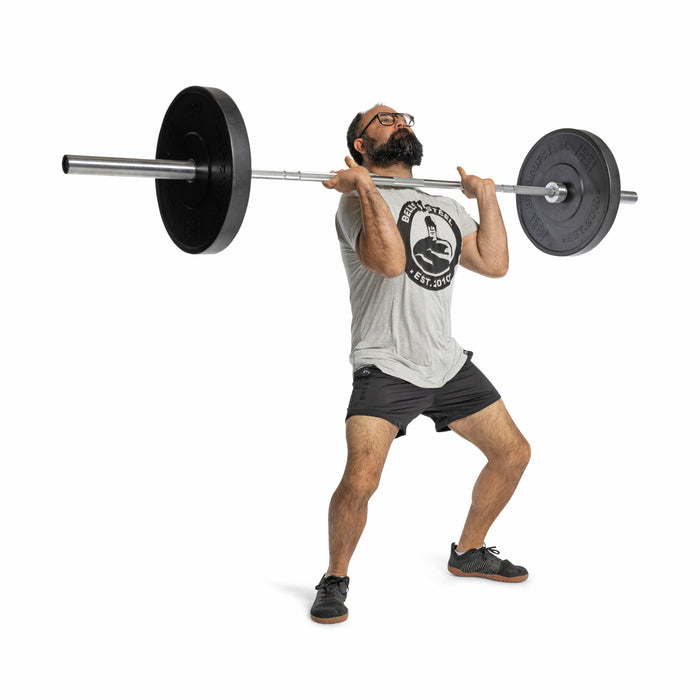 A man in a gray T-shirt, shorts, and glasses lifts a barbell loaded with Bells of Steel All-Black Bumper Plates to his shoulders in a squat stance against a white background.