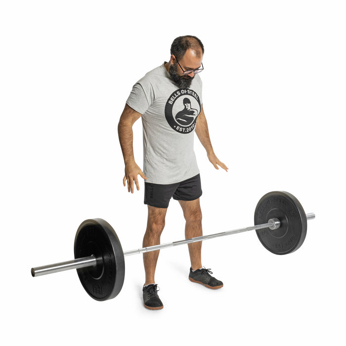 A man in a gray T-shirt and black shorts stands looking down at a barbell loaded with Bells of Steel All-Black Bumper Plates, positioned on the floor in front of him against a white background.
