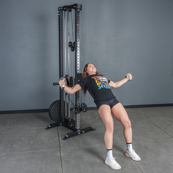 A woman in athletic wear uses the Bells of Steel USA Aluminum Handles Pair with Bearings on a cable machine, leaning back with arms extended, standing on a gray floor against a plain gray wall.