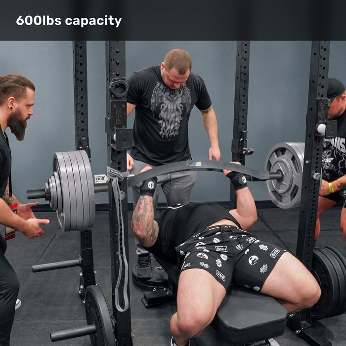 In a gym, a man bench presses a heavily loaded Bells of Steel Arch Nemesis Swiss Bar while three spotters assist. "600lbs capacity" is displayed at the top of the image.