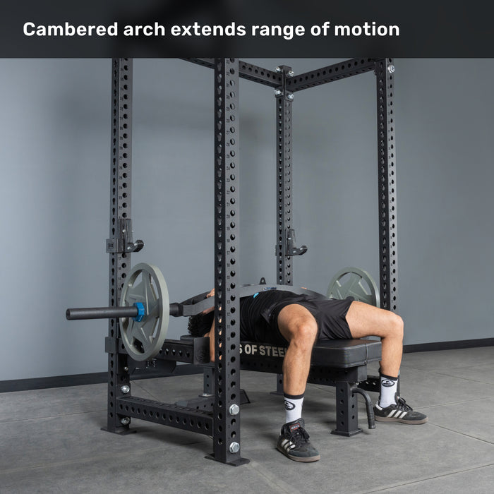 A person uses the Bells of Steel Arch Nemesis Swiss Bar in a power rack, performing a bench press with a cambered arch for greater range of motion. Specialty barbells like this are excellent for boosting your bench. Text: "Cambered arch extends range of motion.