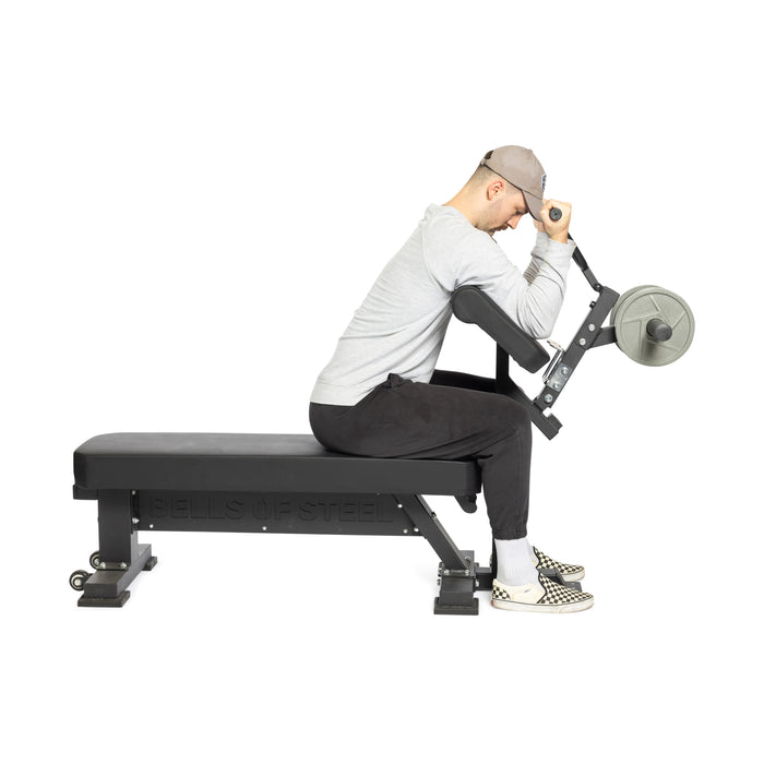 A person uses a preacher curl machine with Bells of Steel Bench Attachments on a Buzzsaw Bench, wearing a light gray cap, gray long-sleeve shirt, black pants, and checkered slip-on shoes against a white background.