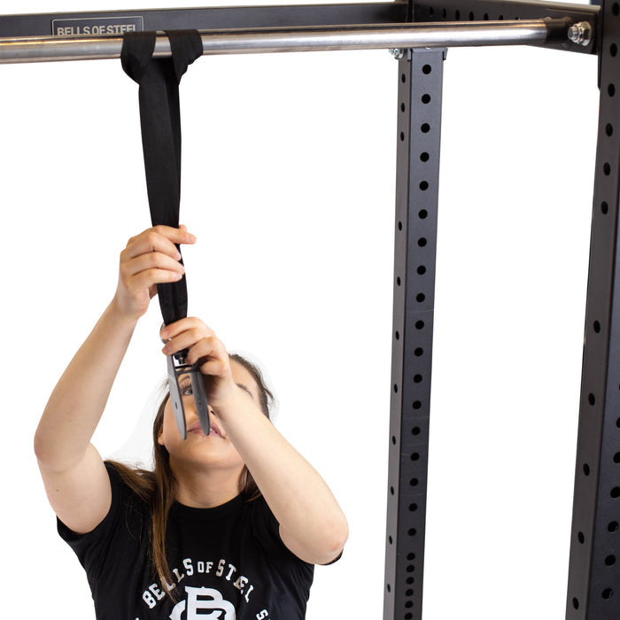 A person attaches a Bells of Steel Cable Pulley hanging strap to a pull-up bar with a metal clasp. They are wearing a black T-shirt and the background is white.