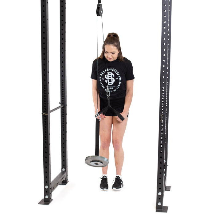 A woman in athletic wear and sneakers stands between two racks, holding a rope attached to the Bells of Steel Cable Pulley, preparing for a forearm workout against a white background.