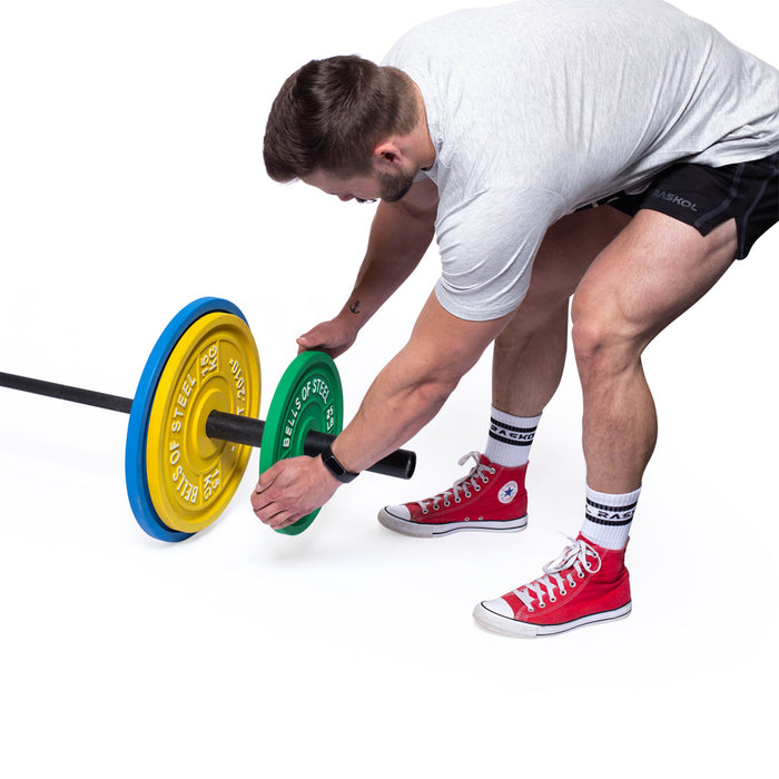 A person wearing a gray T-shirt, black shorts, and red high-tops adjusts Bells of Steel's Calibrated Powerlifting Plates - LB on a barbell against a white backdrop, concentrating on their technique.