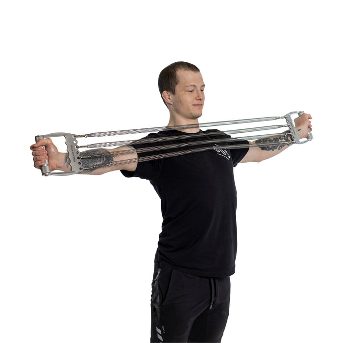 Wearing a black t-shirt and shorts, a tattooed individual uses the Bells of Steel Chest Expander, stretching its springs with arms wide open. Against a plain white background, they embody bodybuilding discipline and focus on muscle building.