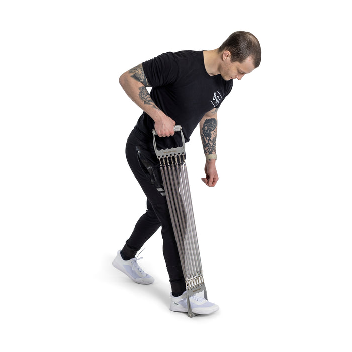 Dressed in black with white shoes and arm tattoos, a person uses the Bells of Steel Chest Expander, pulling resistance bands downward against a plain white backdrop.