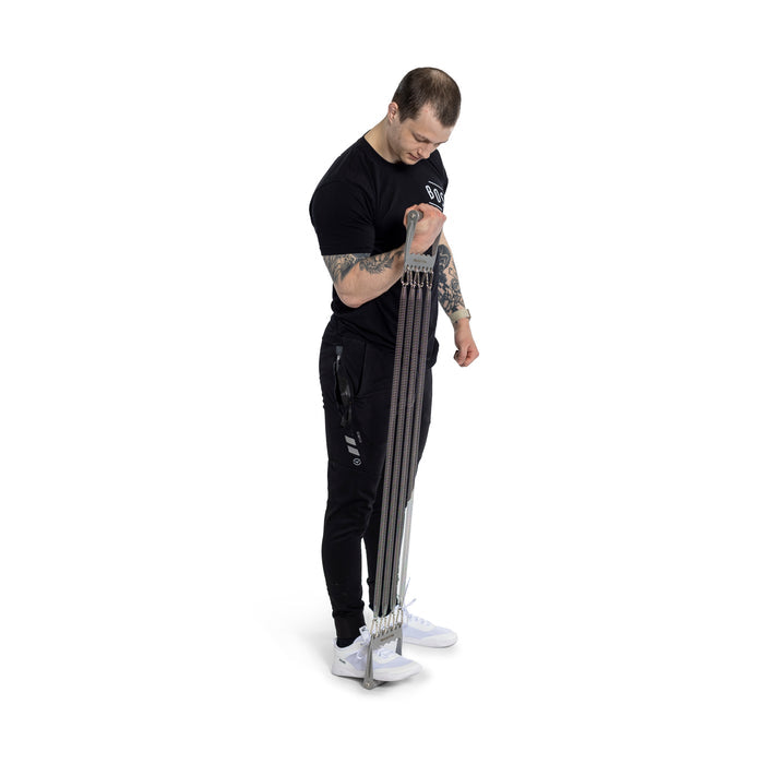 A man in black workout attire uses the Bells of Steel Chest Expander by standing on it and pulling it upwards with one hand, concentrating on bodybuilding against a white, minimalistic background.