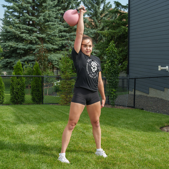 A woman in athletic clothing stands on grass, holding a Bells of Steel Competition Kettlebell overhead with one arm, with trees and a fence in the background.
