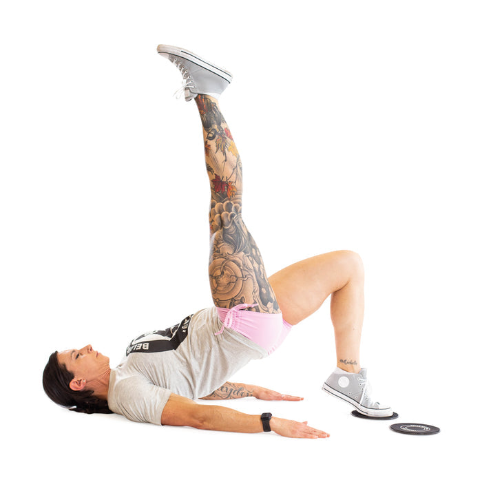 A person in a gray shirt, pink shorts, and sneakers uses Bells of Steel Core Sliders to perform a single-leg glute bridge, extending one leg upward while the other foot rests on the slider for an effective total body workout.