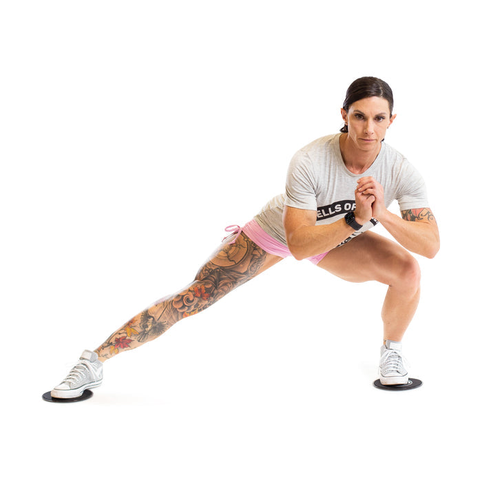 A tattooed woman demonstrates a total body workout with Bells of Steel Core Sliders, performing a lateral lunge by sliding her left leg out while bending her right knee. She wears a gray shirt, pink shorts, and white sneakers on a white background.