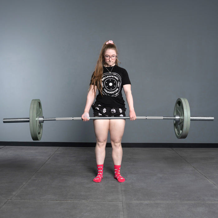 A woman in glasses, a black t-shirt and shorts, and bright pink patterned socks is lifting a Bells of Steel USA Deadlift Bar with weights in a gym with gray walls and flooring.