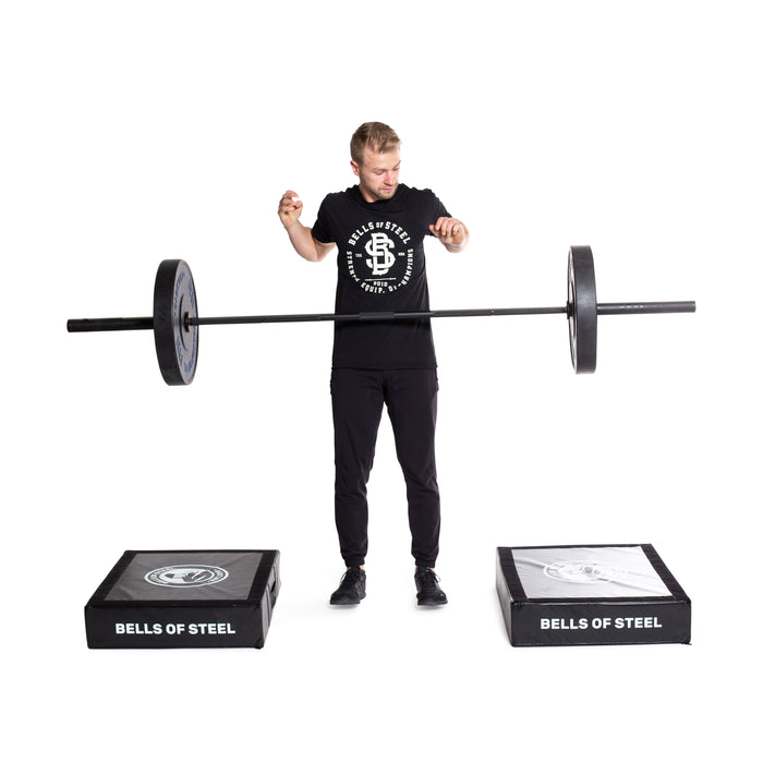 A person in black workout clothes balances a barbell with weights between two Bells of Steel Deadlift Pads (Pair) for noise reduction on a white background.