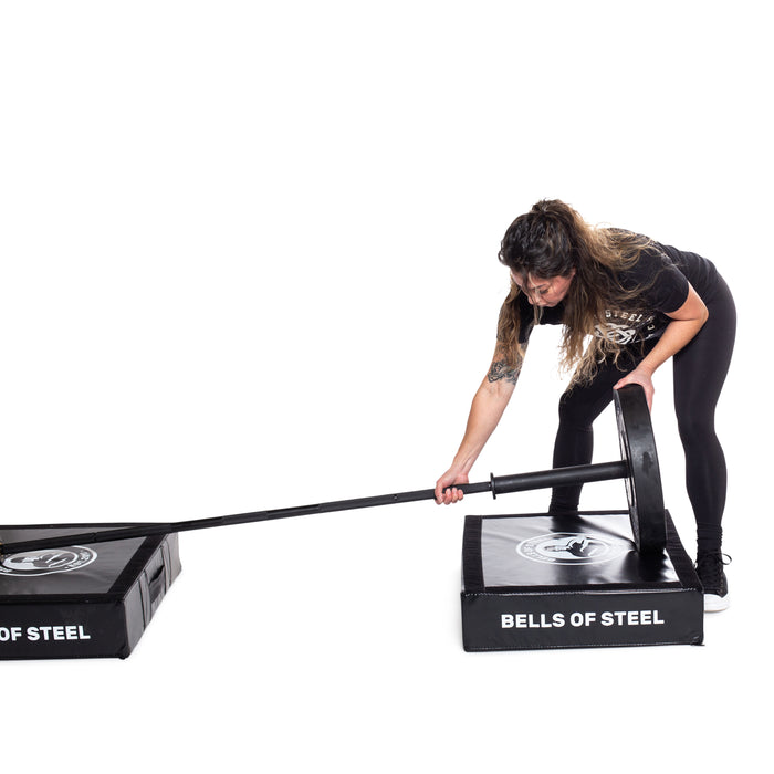 A person loads a weight plate onto a barbell resting between Bells of Steel Deadlift Pads, which reduce weightlifting noise, against a plain white background.