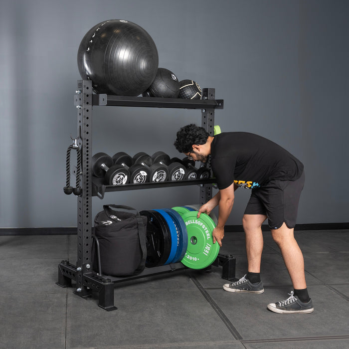A man in athletic wear adds a green weight plate to Bells of Steel USA shelves, organized with Divider for Bumper Plate Shelf (Pack of 3), surrounded by black dumbbells, medicine balls, bands, and gym bags in a gym.