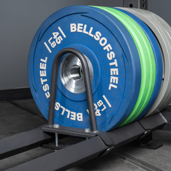 Close-up of blue Bells of Steel USA weight plates on a barbell in a gym, with storage shelves featuring Divider for Bumper Plate Shelf (Pack of 3) visible in the background. The barbell is secured with a black metal holder.