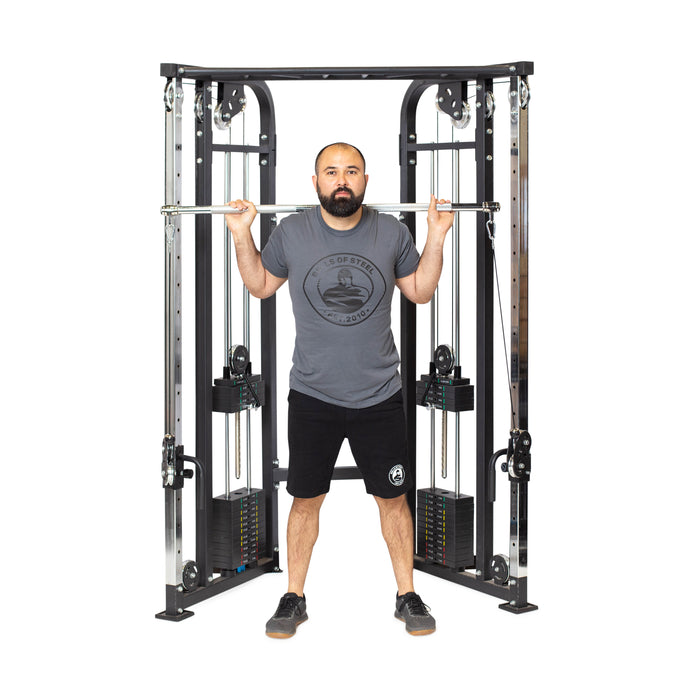 A man stands before a cable machine, gripping the Bells of Steel Double Hook Straight Bar Cable Attachment across his shoulders as he prepares for exercises. He wears a gray shirt, black shorts, and sneakers.