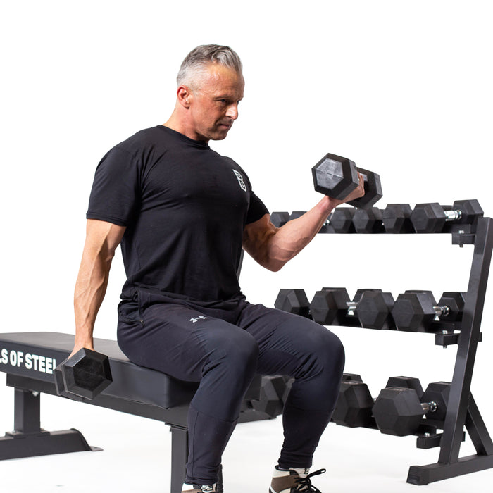 A person in a black t-shirt and pants sits on a bench, performing bicep curls with Bells of Steel's Ergo Rubber Hex Dumbbells. Behind them, bulk sets line the rack, their chrome-plated handles glinting softly against the plain white background.