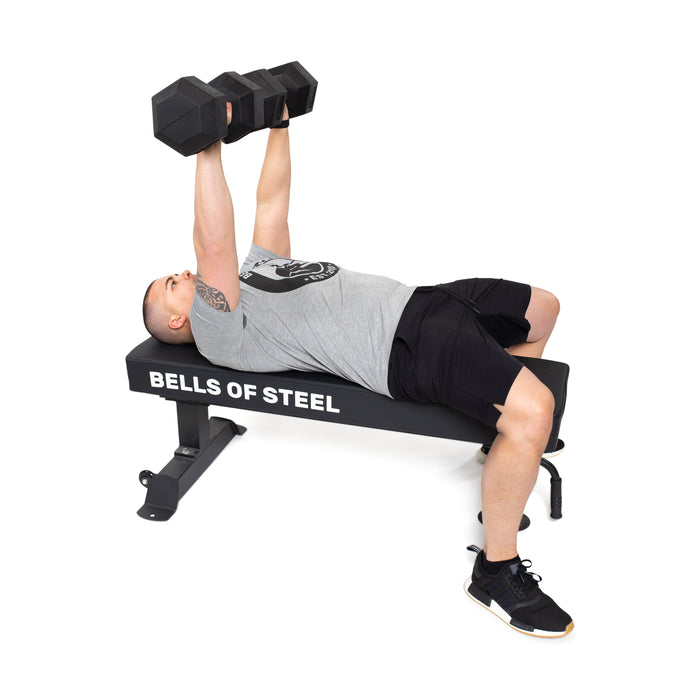 A person wearing a gray shirt, black shorts, and black sneakers with white soles is lying on a weight bench from Bells of Steel. They are doing a bench press with Ergo Rubber Hex Dumbbells that have chrome-plated handles.