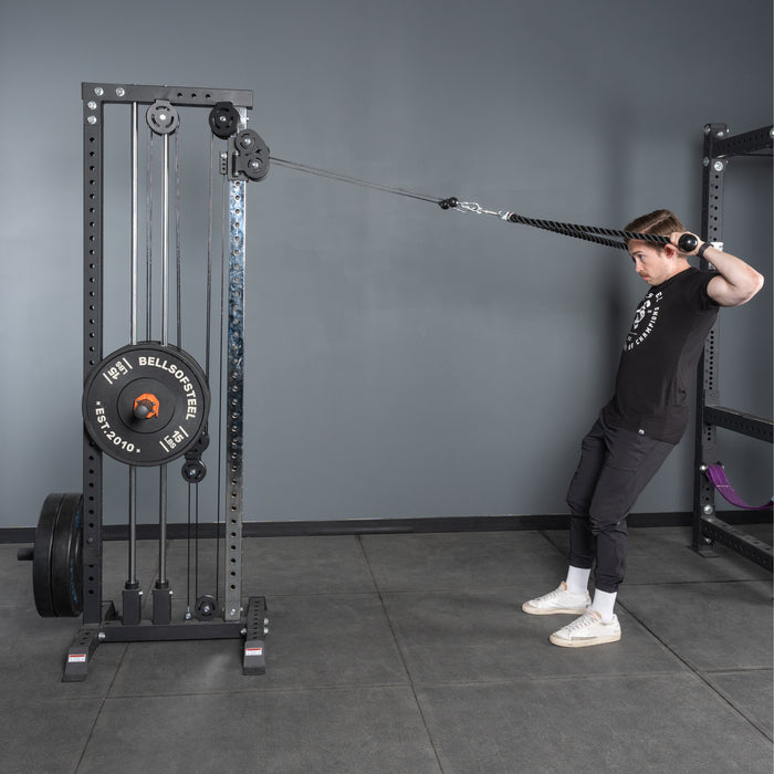 A man in a gym uses the Bells of Steel Extra Long Tricep Rope Extension (137cm) for tricep rope extensions, pulling the rope behind his head while leaning back, engaging his upper back and shoulders. The gym floor and equipment are visible in the background.