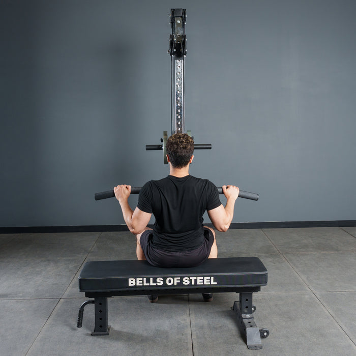 In a gym, a person on a Bells of Steel bench faces a cable machine and grasps the Bells of Steel Fat Bar - Lat Pulldown Bar, ready to perform lat pulldowns to increase grip strength.