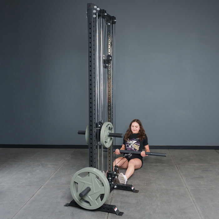 A woman grips the Bells of Steel Fat Bar - Lat Pulldown Bar at a cable machine on the gym floor, preparing for a seated row. The plain gray wall and floor emphasize her grip strength and the bar’s sturdy design.