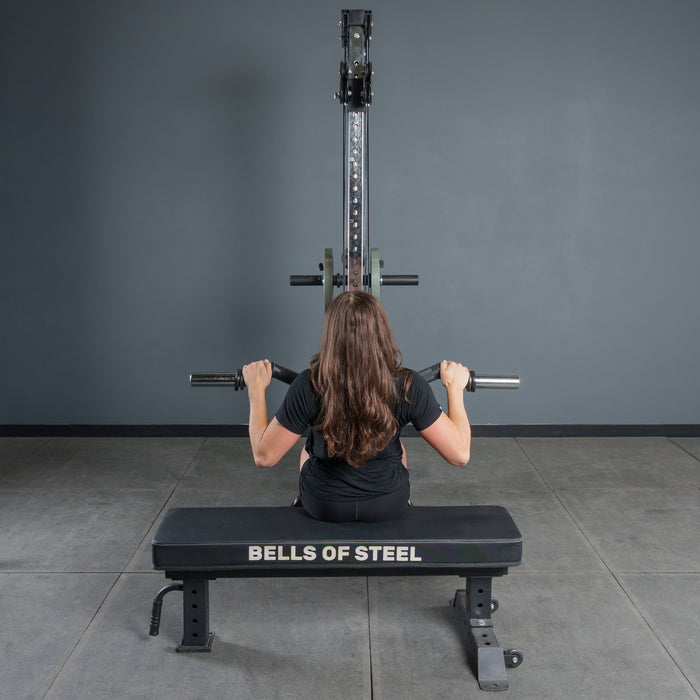 A person with long brown hair sits on a Bells of Steel bench, boosting grip strength while using the Bells of Steel Fat Bar - Olympic Curl Bar Cable Attachment with a lat pulldown, facing a gray wall and gym equipment in the workout room.
