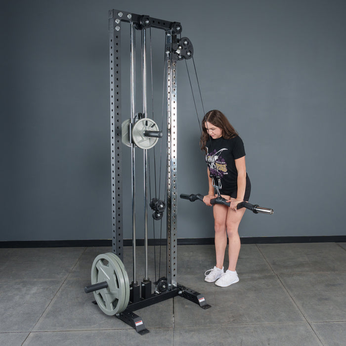 A woman in a black t-shirt and shorts uses the Bells of Steel Fat Bar - Olympic Curl Bar Cable Attachment in a gym, gripping the handle to enhance her grip strength as she faces the equipment on a gray tiled floor.
