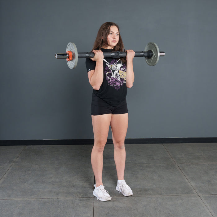 A woman in a black t-shirt and shorts stands indoors, holding the Bells of Steel Fat Bar - Olympic Curl Bar Cable Attachment at shoulder height to enhance grip strength. The background features a plain gray wall and tiled floor.