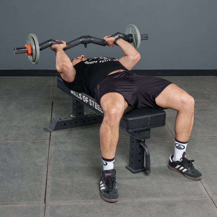 A person lies on a bench press bench, gripping the Bells of Steel Fat Bar - Olympic Curl Bar Cable Attachment above their chest. Wearing black workout gear and white socks, they're ready to test their grip strength.