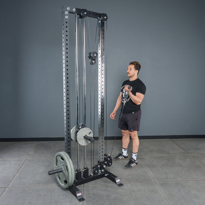 A person stands next to a tall, black cable pulley machine loaded with weight plates, using the Bells of Steel Fat Bar - Single D Handle to improve grip strength in a gym with gray walls and a concrete floor.