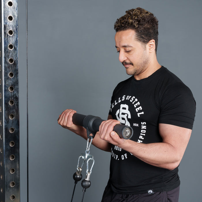 A man in a black Bells of Steel t-shirt does a cable bicep curl in the gym, using the Bells of Steel Fat Bar – Straight Handle Bar attached to resistance cables while looking down at his arms.