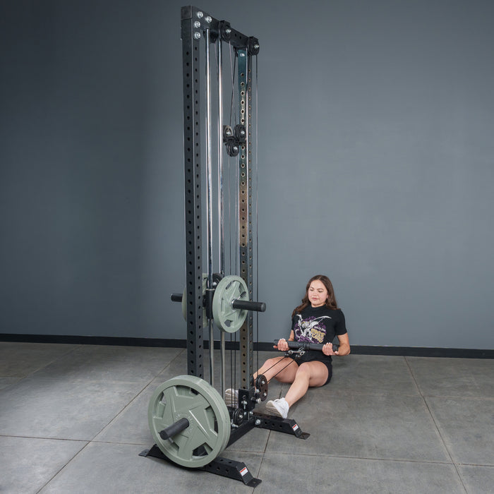 A woman prepares for a seated row in a gym, holding the Bells of Steel Fat Bar - Straight Handle Bar, which is attached to a cable machine with weight plates. The gym features gray walls and a tiled floor.