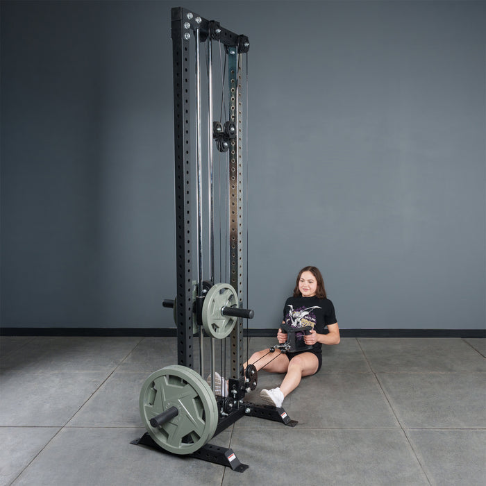 A person sits on the gym floor, performing a seated row with the Bells of Steel Fat Bar - T-Bar Row Cable Attachment on a cable machine with weight plates. The background features a plain gray wall and tiled floor.