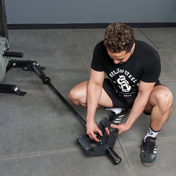 A person in a black Bells of Steel t-shirt and shorts squats on the gym floor, attaching the Bells of Steel Fat Bar - T-Bar Row Cable Attachment to a landmine to boost grip strength for weightlifting.