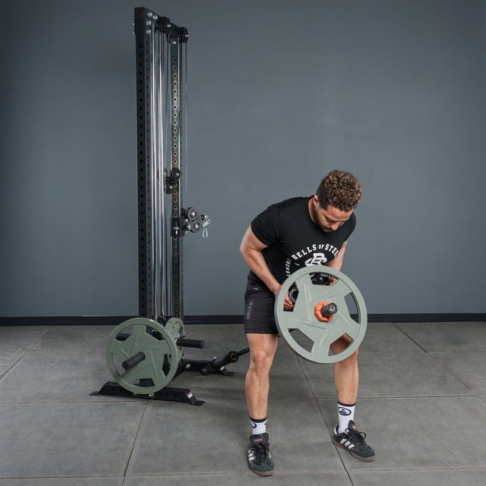A man in a black T-shirt and shorts grips the Bells of Steel Fat Bar - T-Bar Row Cable Attachment by a cable machine in a gym with a plain gray background, highlighting his grip strength as he prepares to lift.