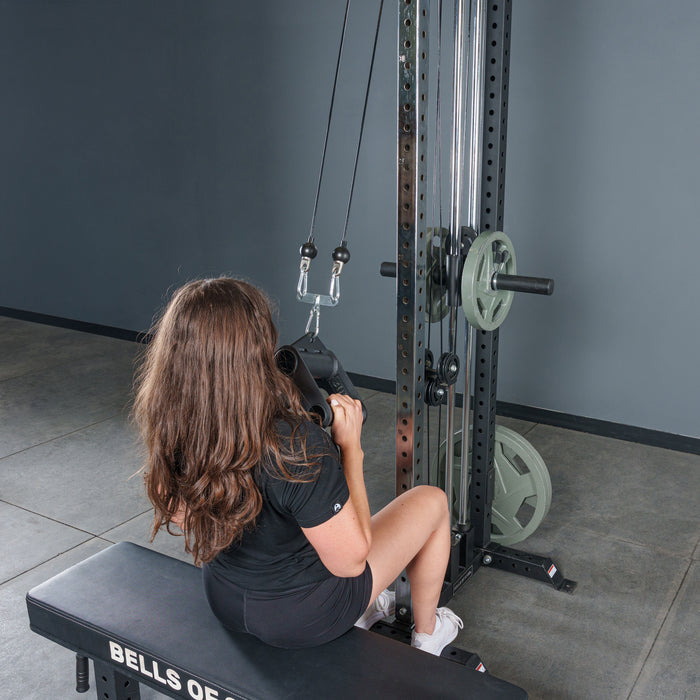 A person with long brown hair, in a black shirt and gray shorts, sits on a “Bells of Steel” bench performing a seated cable row with the Fat Bar - T-Bar Row Cable Attachment to boost grip strength.