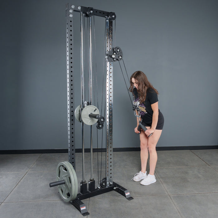 A woman in shorts and a T-shirt stands by a Bells of Steel Fat Bar - Tricep Pressdown machine in the gym, gripping the handle with both hands as she prepares for tricep pushdowns. The machine is loaded with multiple weight plates.