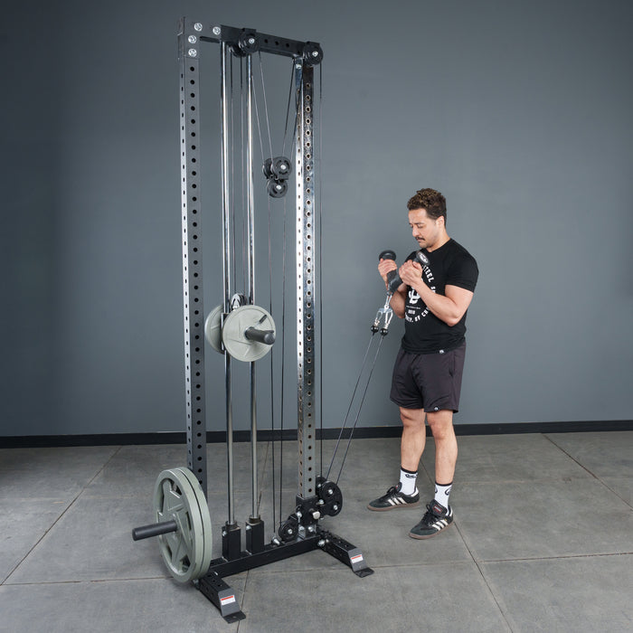 A man in athletic clothing uses the Bells of Steel Fat Bar - Tricep Pressdown on a cable machine in a gym with gray walls and concrete flooring, building grip strength. The machine is loaded with weight plates.