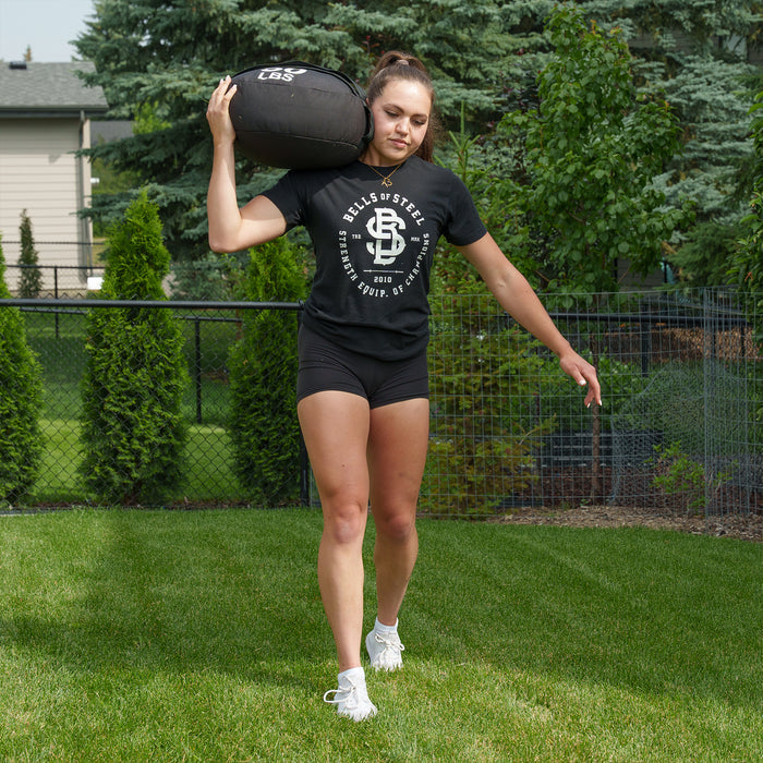 A woman dressed in athletic wear carries a Bells of Steel Fitness Sandbag on her shoulder while walking on grass in a fenced backyard surrounded by trees and shrubs, demonstrating the benefits of sandbag workouts.